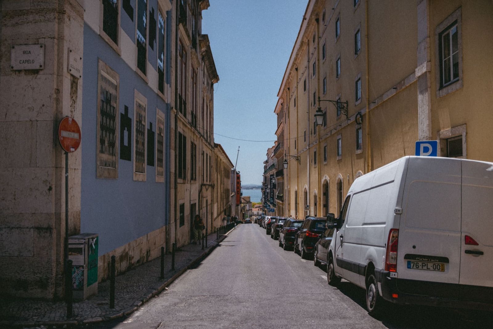 A picturesque street in Lisbon, Portugal, lined with historic buildings and parked vehicles, leading to an ocean view.